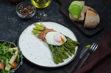 Yeşil kuşkonmaz pastırma ile sarılmış, benedict haşlanmış yumurta, salata ve siyah arka plan üzerinde avokado ile tost. Sağlıklı kahvaltı