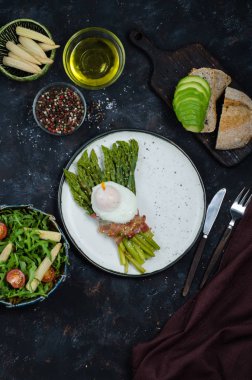 Yeşil kuşkonmaz pastırma ile sarılmış, benedict haşlanmış yumurta, salata ve siyah arka plan üzerinde avokado ile tost. Sağlıklı kahvaltı