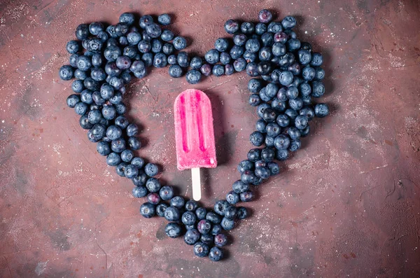 Helado en un palo alrededor de arándanos. Helado casero. Forma de corazón hecha de arándanos sobre un fondo oscuro . — Foto de Stock