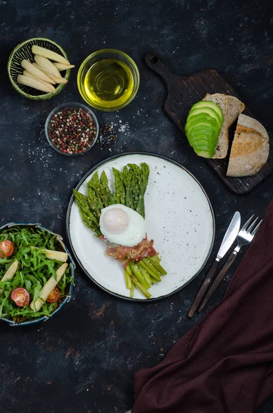 Asparagi verdi avvolti con pancetta, uovo in camicia benedetto, insalata e pane tostato con avocado su sfondo nero. Colazione sana — Foto Stock