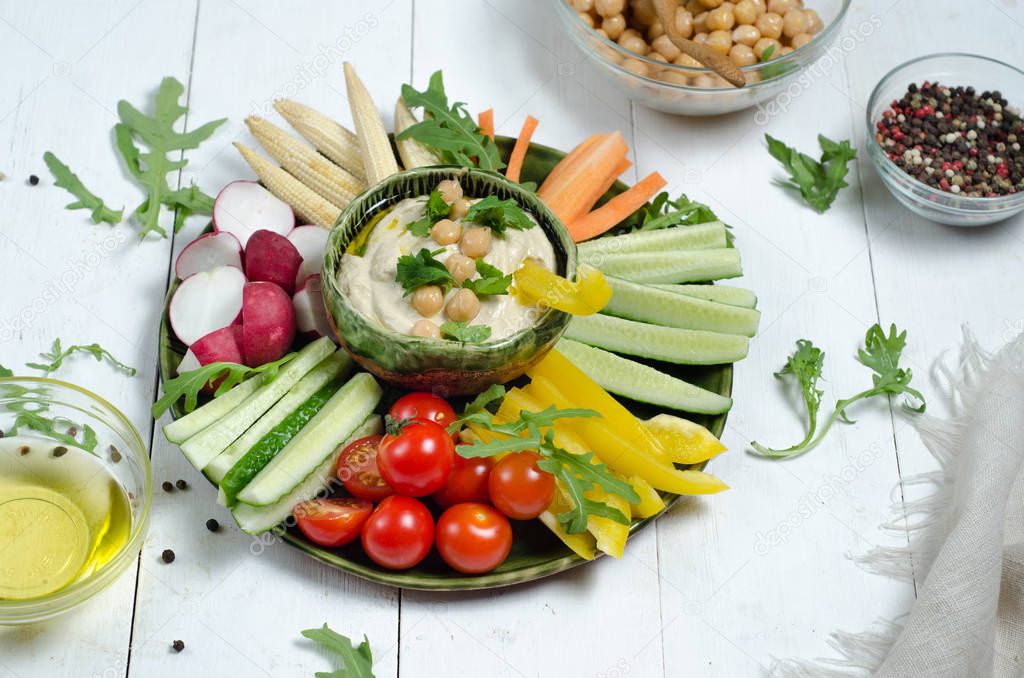 Hummus platter with assorted snacks. Hummus in bowl, vegetables sticks, chickpeas, olive oil. Vegetables, hummus dip,copy space. White wood background