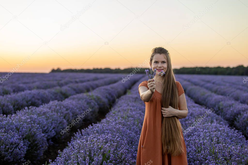 Smiling happy girl in orange dress with long hair holds a lavender bouquet in blooming blossoming beautiful landscape of violet purple lilac lavender field with sunset and orange sky. Summer concept.