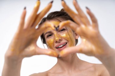 young girl in a cosmetic mask for the face of gold color posing. Shows a different mood. Cheerful, sad, mysterious. Portrait on a blank white background. Makes a heart with your fingers