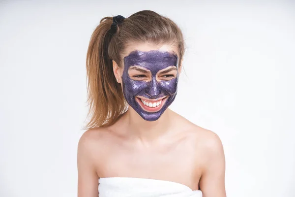 emotional portrait of a young beautiful european girl in violet cosmetic mask on her face. Wrapped in a towel. European, 22 years old, white, tail on the head, blank background, looks away