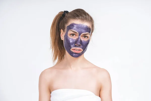 Potret emosional seorang gadis muda european indah dengan topeng kosmetik ungu di wajahnya. Dibungkus dengan handuk. Eropa, 22 tahun, putih, ekor di kepala, latar belakang kosong, melihat lurus — Stok Foto