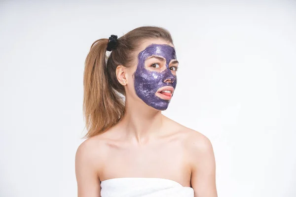 Ritratto emotivo di una giovane bella ragazza europea in maschera cosmetica viola sul suo viso. Avvolto in un asciugamano. Europeo, 22 anni, bianco, coda sulla testa, sfondo bianco, distoglie lo sguardo — Foto Stock