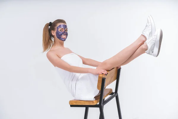 young beautiful naked girl wrapped in a towel in a cosmetic mask on her face sitting on a chair. Sneakers on the legs. Cheerful plot. Blank background, growth portrait