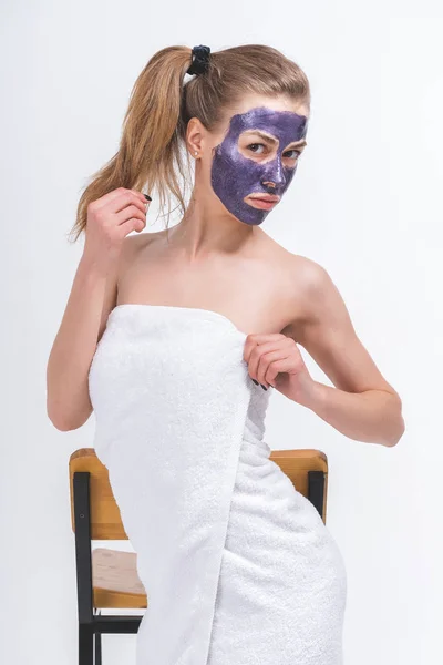 young pretty naked girl wrapped in a towel in a cosmetic mask on her face posing beautifully standing in front of a chair. Half-length portrait on a blank background.