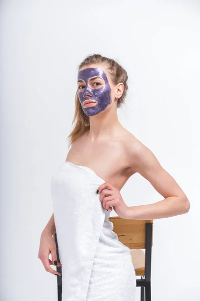 young beautiful naked girl wrapped in a towel in a cosmetic mask on her face posing standing in front of a chair. Half-length portrait on a blank background.