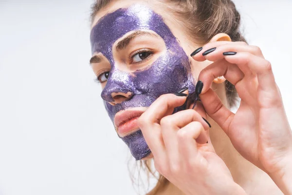 Gadis muda dengan wajah cantik menghilangkan topeng kosmetik ungu. Close-up. Makro . — Stok Foto