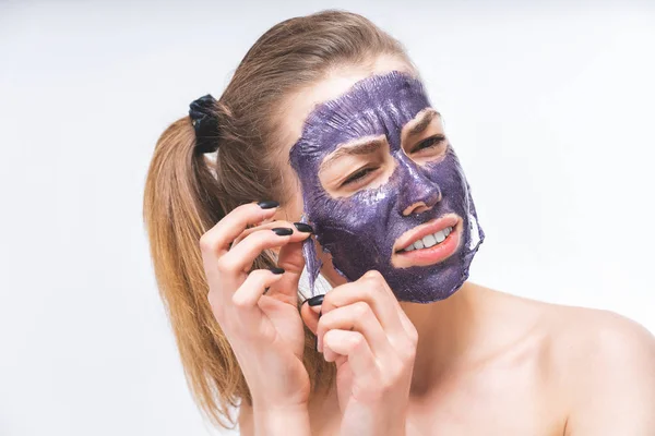 Una giovane ragazza con un bel viso con difficoltà rimuove una maschera cosmetica viola. Primo piano. Macro. Dolore, irritazione. Cura del viso — Foto Stock