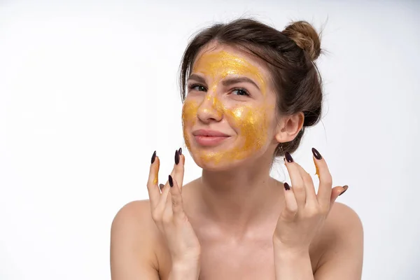 Европейская девушка 25 лет надевает на лицо маску золотого цвета. Увлажняющая маска для лица. В хорошем настроении. Уход за кожей лица . — стоковое фото