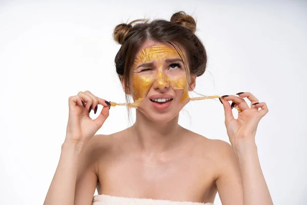 A young girl, 25 years old, removes a cosmetic mask from her face. Golden color. Hands in the frame. Large emotional portrait. Tears off the flaps. Different mood. — Stock Photo, Image