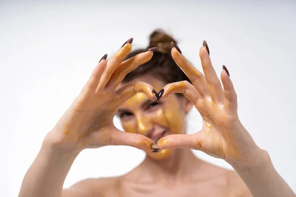 Gadis muda dalam topeng kosmetik untuk wajah berpose warna emas. Menunjukkan suasana hati yang berbeda. Ceria, sedih, misterius. Potret pada latar belakang putih kosong. Membuat hati dengan jari-jarimu — Stok Foto