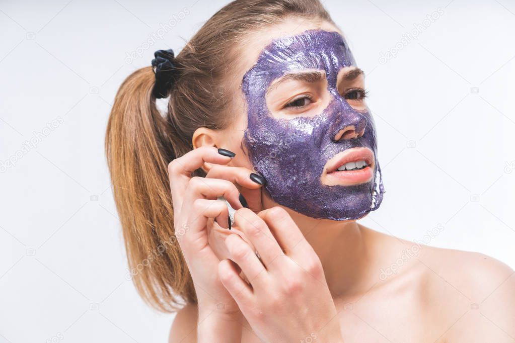 a young girl with a beautiful face with difficulty removes a purple cosmetic mask. Close-up. Macro. Pain, irritation. Facial care