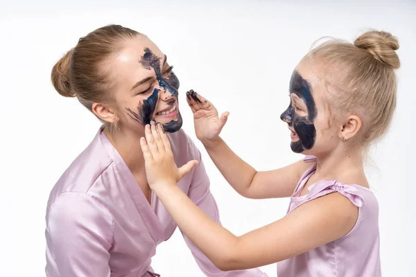 Petite fille fait de sa mère un masque cosmétique sur son visage. La fille se soucie, aide maman. Petite fille et jeune mère . — Photo
