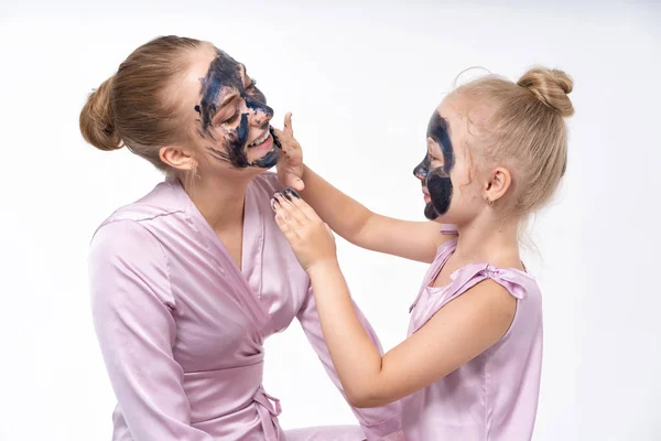 La figlioletta fa di sua madre una maschera cosmetica sul viso. Alla figlia importa, aiuta la mamma. Figlia piccola e giovane madre . — Foto Stock