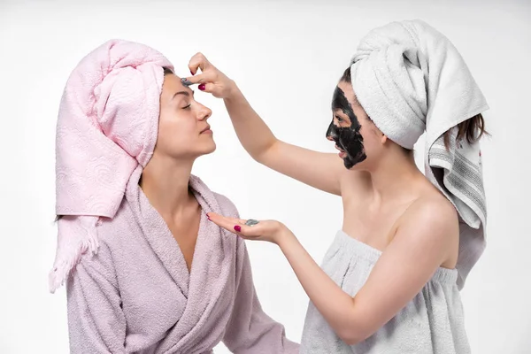 A menina coloca sua irmã uma máscara cosmética em seu rosto. Eles se divertem e se entregam ao processo. Antecedentes vazios, bom humor. Cuidado com a pele facial. Irmãs . — Fotografia de Stock