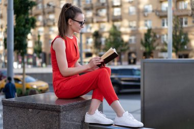 kırmızı takım elbise li ve gözlüklü genç ve şık bir Avrupalı kız sokaktaki parapetin üzerine oturur ve kitap okur. Onun insanlarının geçişini geçti, şehir hayatını görebilirsin. Küçük bir imza için bir yöntem vardır.