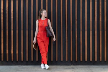 kırmızı takım elbiseli ince genç kız ahşap kirişler ile duvara yakın duruyor. Elinde bir defter ve bir çanta var. Dinlenmek ve programı hatırlamak için kalktı. Kopyalama alanı