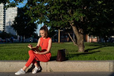 Avrupalı kız 25 yaşında kırmızı takım elbiseli bir şehir meydanında kaldırımda oturan ve coşkuile bir kitap okuma, büyük bir mola sahip, dinlenme. Kopyalama alanı için harika fırsatlar