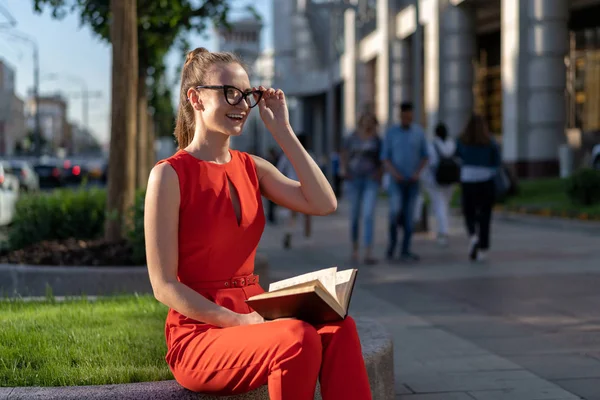 Una joven europea con estilo en un traje rojo y gafas se sienta en el parapeto en la calle y lee un libro. Más allá de que su gente pase, se puede ver la vida de la ciudad. Hay un método para una pequeña firma . — Foto de Stock