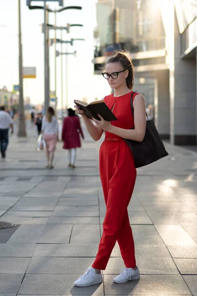 Una joven europea con estilo en un traje rojo y gafas se sienta en el parapeto en la calle y lee un libro. Más allá de que su gente pase, se puede ver la vida de la ciudad. Hay un método para una pequeña firma . — Foto de Stock