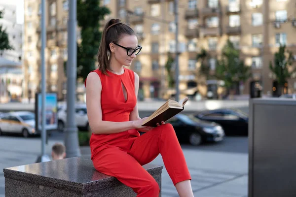 Una joven europea vestida con un traje rojo y gafas está sentada en el parapeto cerca de la acera y la calzada y leyendo un libro, mirando un cuaderno. Copiar espacio . — Foto de Stock