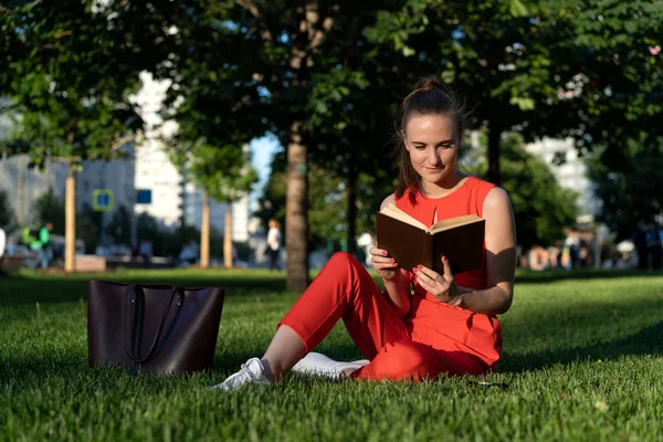 Chica europea de 25 años con un traje rojo sentada en el césped en una plaza de la ciudad y leyendo un libro con entusiasmo, teniendo un descanso. Junto a ella hay una gran bolsa marrón. Gran oportunidad para copiar espacio — Foto de Stock