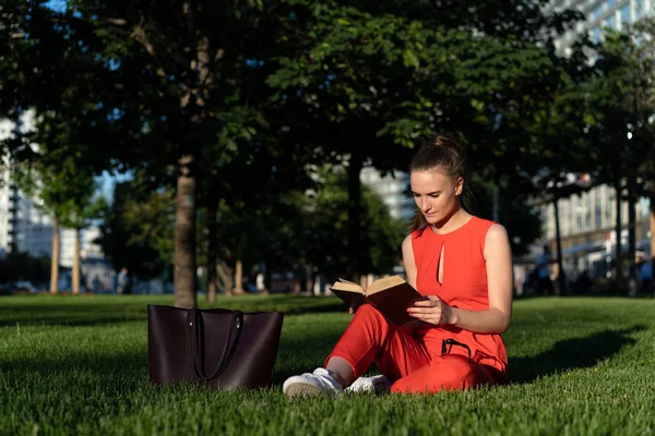 Chica europea de 25 años con un traje rojo sentada en el césped en una plaza de la ciudad y leyendo un libro con entusiasmo, teniendo un descanso. Junto a ella hay una gran bolsa marrón. Gran oportunidad para copiar espacio —  Fotos de Stock