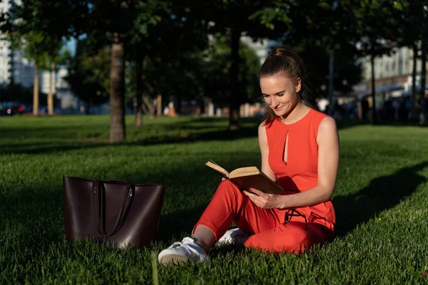 Chica europea de 25 años con un traje rojo sentada en el césped en una plaza de la ciudad y leyendo un libro con entusiasmo, teniendo un descanso. Junto a ella hay una gran bolsa marrón. Gran oportunidad para copiar espacio —  Fotos de Stock