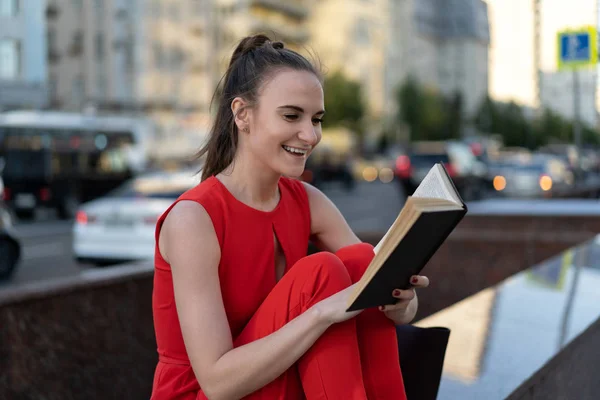 Niña con un traje rojo y zapatillas blancas se sienta en el parapeto con un cuaderno, un libro en sus manos. Hermosa vista del tráfico de la ciudad. Buenas noches, puesta de sol. Copiar espacio — Foto de Stock