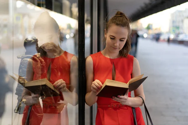 Chica se para cerca de la pared de cristal y mira en el libro, portátil. Uno puede ver su reflejo en la luz de la tarde. Tranquilo y hermoso europeo . — Foto de Stock