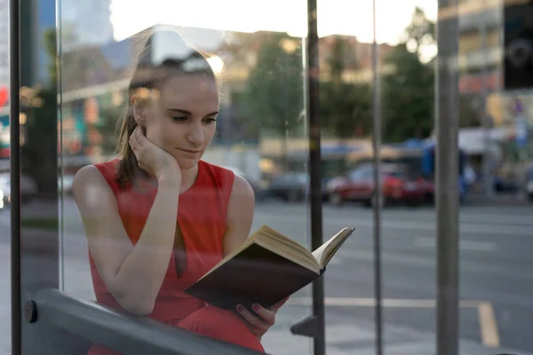 Молода вродлива дівчина з книжкою в руках сидить на автобусній зупинці. Вигляд крізь скло, приємна вечірня атмосфера спокою. Копіюю простір. Спосіб життя — стокове фото