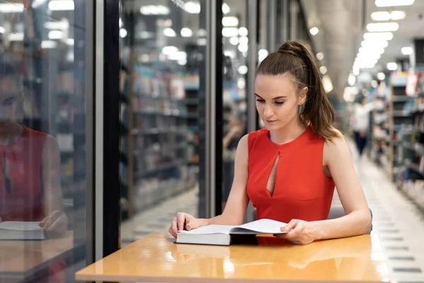 Una joven blanca con un traje rojo se sienta en una mesa en la biblioteca, una librería, una oficina y mira un cuaderno, lee un libro. Hermosa perspectiva. Reflexiones. espacio de copia — Foto de Stock