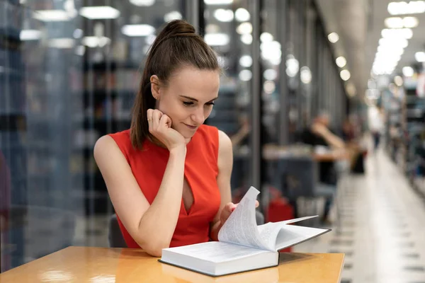 Молода біла дівчина в червоному костюмі сидить за столом у бібліотеці, книгарні, кабінеті і дивиться на блокнот, читає книгу. Прекрасна перспектива. Роздуми. копіювання простору — стокове фото