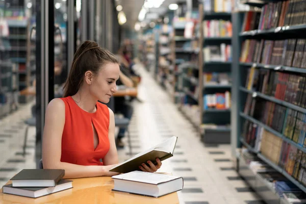Una joven blanca con un traje rojo se sienta en una mesa en la biblioteca, una librería, una oficina y mira un cuaderno, lee un libro. Hermosa perspectiva. Reflexiones. espacio de copia — Foto de Stock