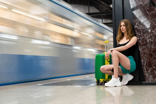 Eine schöne junge weiße Europäerin steht mit einem grünen Koffer an der U-Bahn-Station und wartet auf den Zug. Themenreise. — Stockfoto