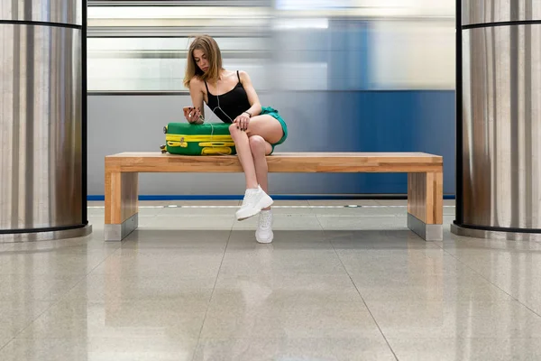 European girl sitting on a bench with a phone in her hands and in the headphones, next to a travel suitcase. It is located at the railway station or subway station. Theme of independent travel. — Stock Photo, Image