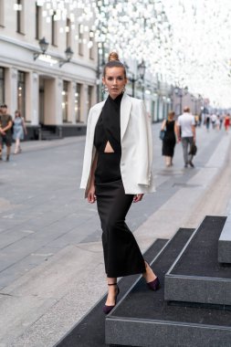 Siyah uzun elbiseli ve beyaz ceketli genç kız boş bir caddenin arka planında poz veriyor. Tam yükseklik. Çok şık görünüyorsun.