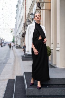 Siyah uzun elbiseli ve beyaz ceketli genç kız boş bir caddenin arka planında poz veriyor. Tam yükseklik. Çok şık görünüyorsun.