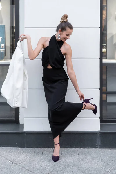 Piękna biała stylowa dziewczyna w czarnej sukni trzyma w ręku białą kurtkę i pierwotnie stwarzając na tle sklepu mody — Zdjęcie stockowe