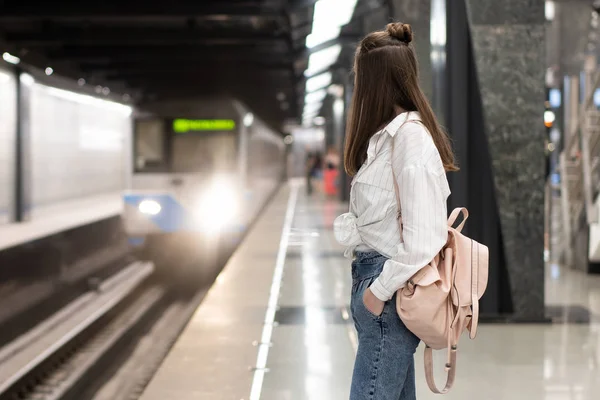 Pantolon ve sırt çantalı genç Avrupalı kız metro istasyonunda treni bekliyor. Hareketli arka plan, canlı bir atmosfer iletmek için bulanık. Bu harika bir fikir..