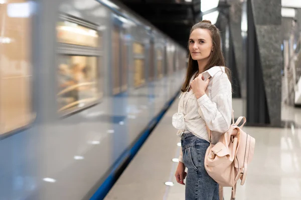 young white girl with long hair. With a backpack. Standing at the metro station and waiting for the train. The train is out of focus to convey the effect of the reality of the movement.
