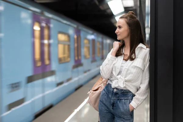 Joven chica blanca con el pelo largo. Con una mochila. De pie en la estación de metro y esperando el tren. El tren está fuera de foco para transmitir el efecto de la realidad del movimiento . — Foto de Stock