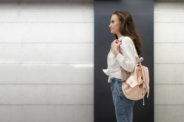 European girl with a backpack, in jeans and shirt stands against the background of an empty wall and looks around. The usual everyday situation. Copy space