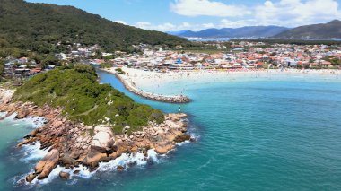 Florianpolis Santa Catarina Brezilya 'daki Barra da Lagoa Plajı' nın panoramik hava manzarası güneşli bir yaz gününde turkuaz mavi deniz ve kayalık kıyıları ve bol Atlantik ormanlarıyla dolu kalabalık bir plaj.