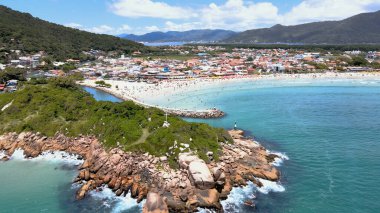 Florianpolis Santa Catarina Brezilya 'daki Barra da Lagoa Plajı' nın panoramik hava manzarası güneşli bir yaz gününde turkuaz mavi deniz ve kayalık kıyıları ve bol Atlantik ormanlarıyla dolu kalabalık bir plaj.