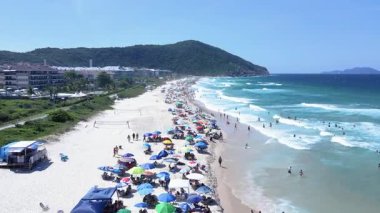Brava sahilinin insansız hava aracı görüntüsü, kum ve denizin üzerinde uçan, birçok insan şemsiyeli ve deniz kenarındaki çeşitli binalar ve evler, Florianopolis, Santa Catarina, Brezilya.