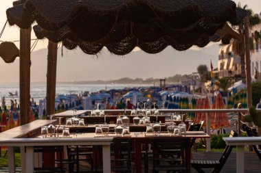Açık İtalyan kafe veya restoranındaki masalar gün batımında deniz manzaralı kalabalık kumlu plajda akşam yemeği için servis ediliyor.